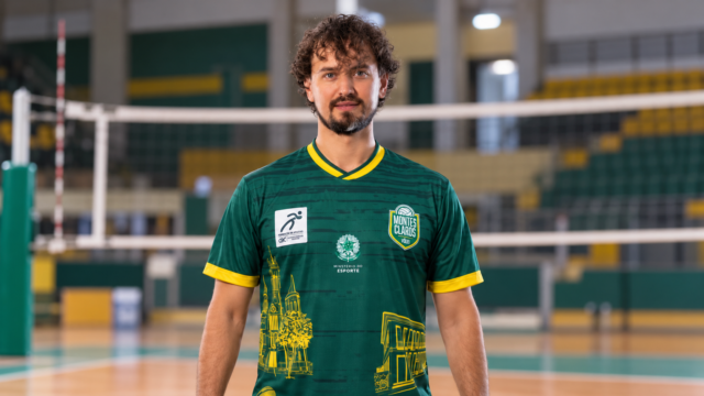 Vitinho Hillmann com uniforme verde do MOC Vôlei, em composição com ginásio de vôlei ao fundo.